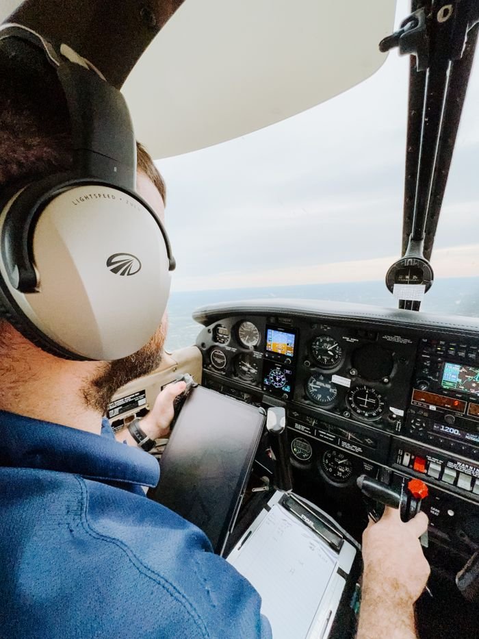 Pilot operating a small aircraft from the cockpit, wearing headphones and navigating at sunset.